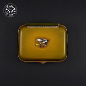 Może przedstawiać: Prostokątne, oliwkowozielone skórzane pudełko do łowienia muchowego ze szczegółowym wzorem przynęty. Pudełko ma zawias ze skóry w kolorze ciemnozielonym i małe metalowe zapięcie. Na górze pudełka znajduje się przynęta z białymi i czarnymi paskami, szarym korpusem oraz zielonym i pomarańczowym haczykiem.