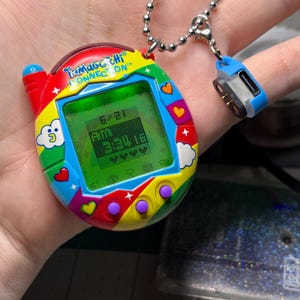 May include: A Tamagotchi Connection digital pet device featuring a vibrant rainbow-colored shell with cloud, heart, and star graphics. The screen displays the date 6/21 and time 3:34. A silver ball chain is attached to a small blue and black connector.