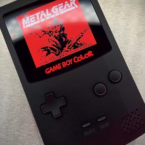 May include: A black Game Boy Color handheld console displaying the "Metal Gear Solid" game on its screen. The screen shows a red background with white text and a character graphic. The console has a directional pad, two action buttons, and "SELECT" and "START" buttons.