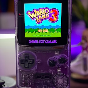 May include: A transparent purple Game Boy Color handheld console displaying the "Wario Land 3" game. The screen shows a colorful game scene with text. The console has a power indicator, directional pad, and action buttons. The text "GAME BOY COLOR" is visible.