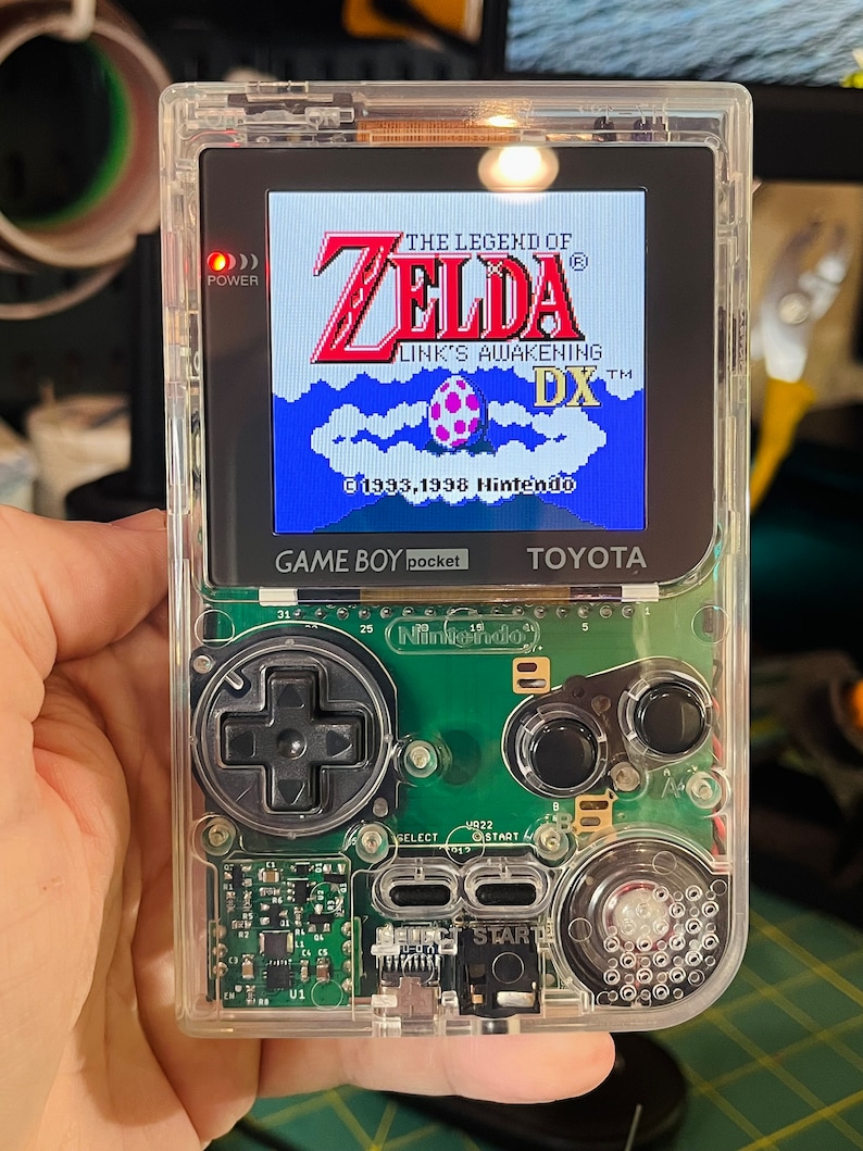 May include: A clear Game Boy Color handheld gaming console with a green circuit board. The screen displays the game "The Legend of Zelda: Link's Awakening DX" with the title in red and the game logo in blue. The power button is lit red.