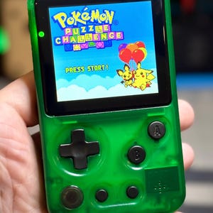 May include: A handheld video game console with a translucent green plastic shell. The screen displays the title Pokémon Puzzle Challenge with Pikachu and Pichu holding balloons. It features a black directional pad, A and B buttons, and small control buttons.