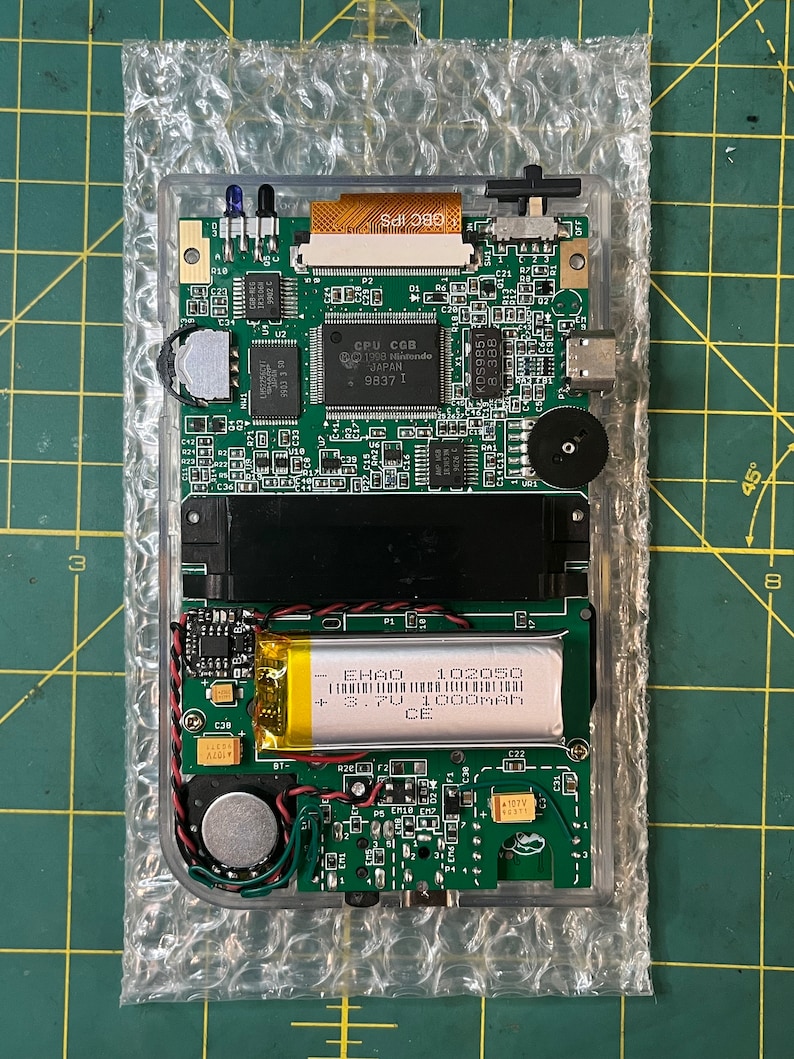May include: Close-up of a green circuit board with a black and silver battery labeled "EHAO 102050 + 3.7V 1000mAh CE". The board has several components, including a small speaker, a switch, and a connector.