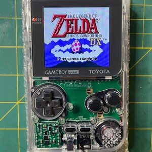 May include: A clear Game Boy Color handheld gaming console with a green circuit board and a screen displaying the game "The Legend of Zelda: Link's Awakening DX". The game's title screen shows Link standing in front of a blue sky with white clouds and a pink egg.