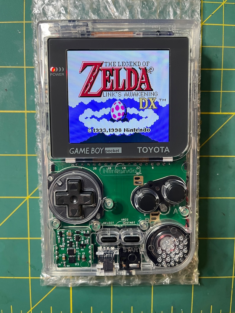 May include: A clear Game Boy Color handheld gaming console with a green circuit board and a screen displaying the game "The Legend of Zelda: Link's Awakening DX". The game's title screen shows Link standing in front of a blue sky with white clouds and a pink egg.