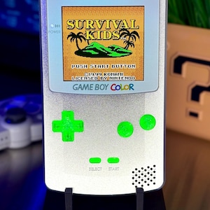 May include: A silver Game Boy Color console replica with a screen displaying the "Survival Kids" game. The console has green buttons and a black stand. The text "GAME BOY COLOR" is visible on the console and stand.