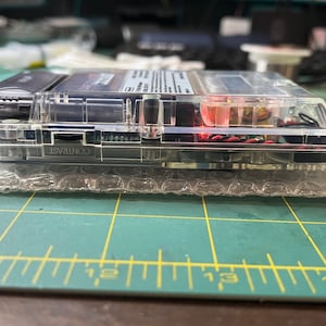 May include: A clear, transparent handheld gaming device with a black and silver cartridge inserted. The device has a contrast knob and is resting on a bubble wrap surface.