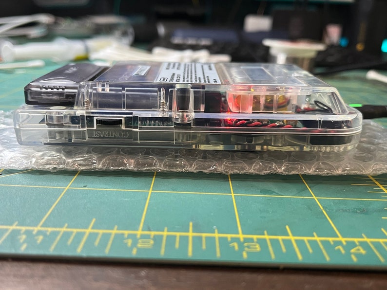 May include: A clear, transparent handheld gaming device with a black and silver cartridge inserted. The device has a contrast knob and is resting on a bubble wrap surface.