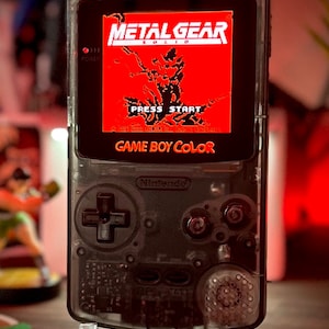May include: A clear Game Boy Color handheld gaming console with a translucent black shell. The console is displaying the title screen for the game Metal Gear Solid. The screen is red with black text that reads "METAL GEAR SOLID" and "PRESS START". The text "GAME BOY COLOR" is displayed in white on a black background.