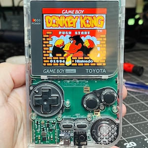 May include: A clear, transparent Game Boy Color handheld gaming console with a Donkey Kong game cartridge inserted. The screen displays the game title "Donkey Kong" and the text "Push Start" in red and yellow. The console has a green circuit board with buttons and a black D-pad.