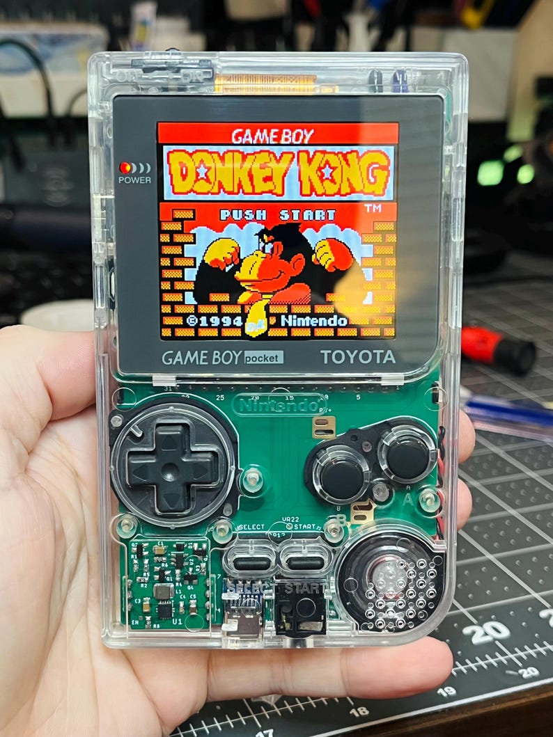 May include: A clear, transparent Game Boy Color handheld gaming console with a Donkey Kong game cartridge inserted. The screen displays the game title "Donkey Kong" and the text "Push Start" in red and yellow. The console has a green circuit board with buttons and a black D-pad.