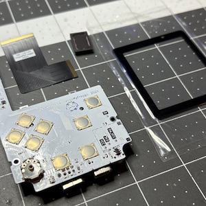 May include: A white circuit board with gold-colored buttons and a black ribbon cable. The board has text that reads "Leo My Froggo 2024".