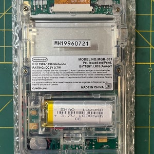 May include: A clear plastic Game Boy Color handheld gaming console with a silver faceplate. The console has a model number MH19960721, a battery compartment, and a label that reads "MODEL NO. MGB-001 Pat. Issued and Pend. BATTERY: LR03 (AAA)x2". The console is made in China.