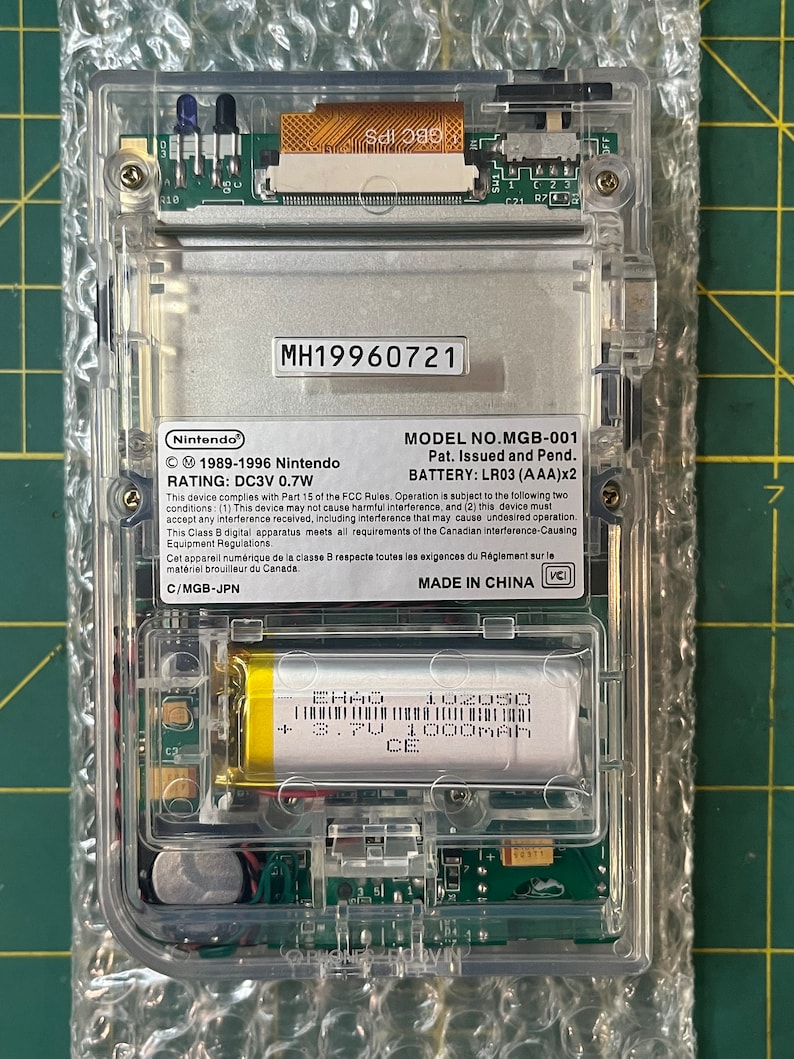 May include: A clear plastic Game Boy Color handheld gaming console with a silver faceplate. The console has a model number MH19960721, a battery compartment, and a label that reads "MODEL NO. MGB-001 Pat. Issued and Pend. BATTERY: LR03 (AAA)x2". The console is made in China.