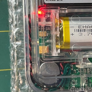 May include: Close-up of a clear plastic case with a red LED light, a circuit board, a battery, and a silver component. The case has the text "This Class B digital apparatus meets all requirements. Equipment Regulations. Cet appareil num&eacute;rique de la classe B respecte mat&eacute;riel brouilleur du Canada. C/MGB-JPN" printed on it.