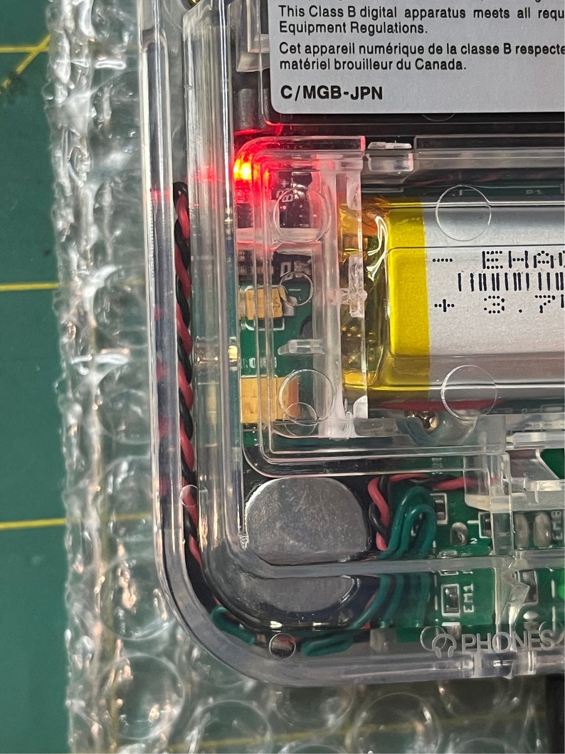 May include: Close-up of a clear plastic case with a red LED light, a circuit board, a battery, and a silver component. The case has the text "This Class B digital apparatus meets all requirements. Equipment Regulations. Cet appareil num&eacute;rique de la classe B respecte mat&eacute;riel brouilleur du Canada. C/MGB-JPN" printed on it.