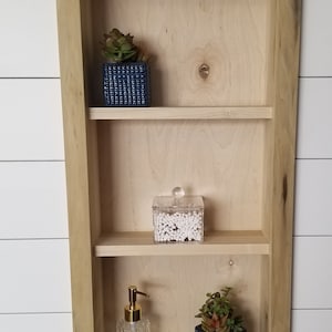 Pu&ograve; includere: Mensola da parete in legno chiaro a tre livelli, con una piccola succulenta in un vaso quadrato blu, un contenitore trasparente di cotton fioc e un dispenser di sapone decorativo.