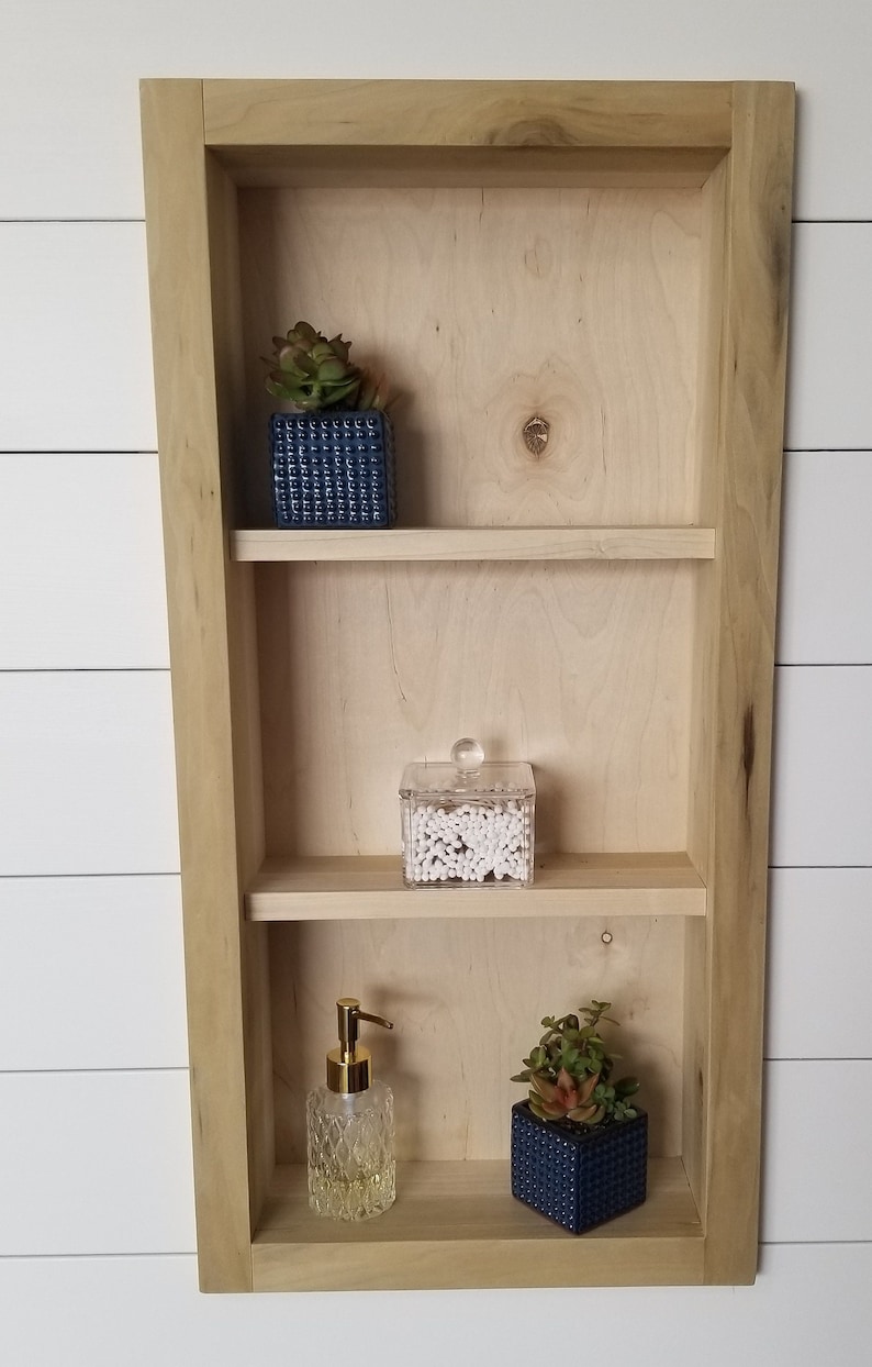 Pu&ograve; includere: Mensola da parete in legno chiaro a tre livelli, con una piccola succulenta in un vaso quadrato blu, un contenitore trasparente di cotton fioc e un dispenser di sapone decorativo.