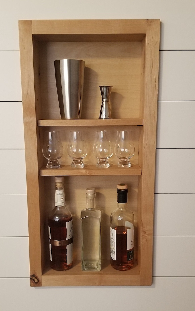 Pu&ograve; includere: Una mensola da bar in legno con uno shaker per cocktail in acciaio inossidabile, un jigger doppio, sei bicchieri e tre bottiglie di liquore. La mensola &egrave; montata su una parete bianca.