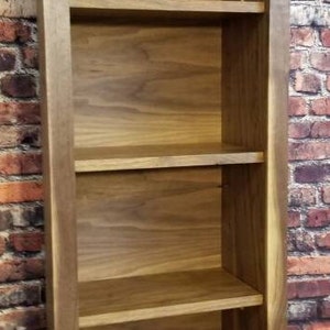 Pu&ograve; includere: Una libreria alta in legno con quattro ripiani. La libreria &egrave; realizzata in legno marrone chiaro e ha un design semplice e rettangolare. I ripiani sono distanziati uniformemente e sembrano robusti. La libreria &egrave; contro un muro di mattoni.