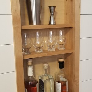 Pu&ograve; includere: Un mobile bar in legno incassato in una parete, con uno shaker per cocktail in acciaio inossidabile, un jigger, sei bicchieri a stelo e tre bottiglie di liquore. Il mobile &egrave; in legno chiaro ed &egrave; montato su una parete bianca.