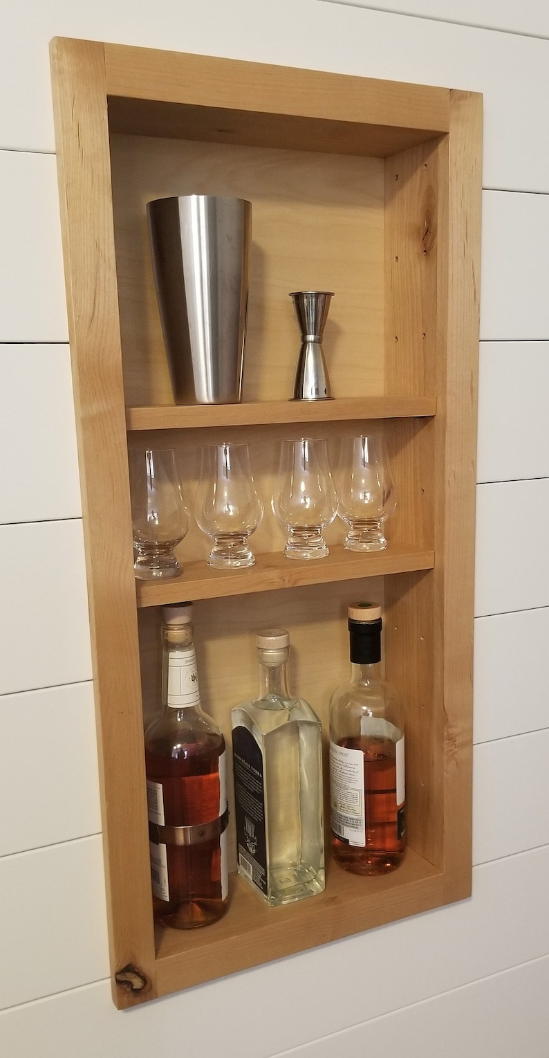 Pu&ograve; includere: Un mobile bar in legno incassato in una parete, con uno shaker per cocktail in acciaio inossidabile, un jigger, sei bicchieri a stelo e tre bottiglie di liquore. Il mobile &egrave; in legno chiaro ed &egrave; montato su una parete bianca.