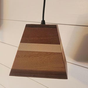 Op de afbeelding: Een houten hanglamp met een geometrische, piramidevormige kap. De kap is gemaakt van meerdere lagen hout, elk met een ander nerfpatroon en kleur, wat een unieke en rustieke uitstraling creëert.