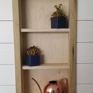 Pu&ograve; includere: Una mensola da parete in legno a tre livelli, con due piccole piante grasse in vaso in vasi quadrati blu e un annaffiatoio in rame. La mensola &egrave; in legno chiaro ed &egrave; montata su una parete a doghe bianche.