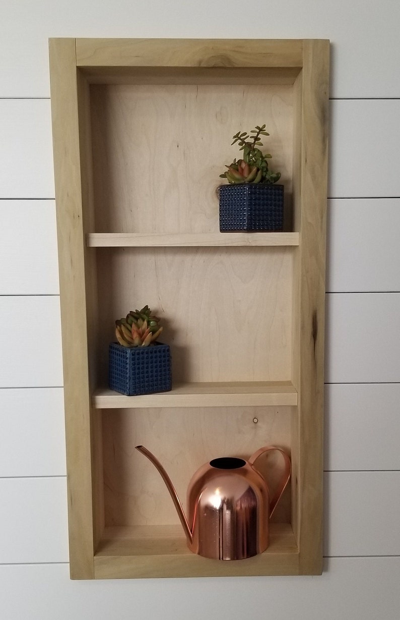Pu&ograve; includere: Una mensola da parete in legno a tre livelli, con due piccole piante grasse in vaso in vasi quadrati blu e un annaffiatoio in rame. La mensola &egrave; in legno chiaro ed &egrave; montata su una parete a doghe bianche.