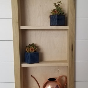 Pu&ograve; includere: Una mensola da parete in legno a tre livelli, con due piccoli vasi quadrati blu con succulente e un annaffiatoio in rame. La mensola &egrave; in legno chiaro ed &egrave; montata su una parete bianca.