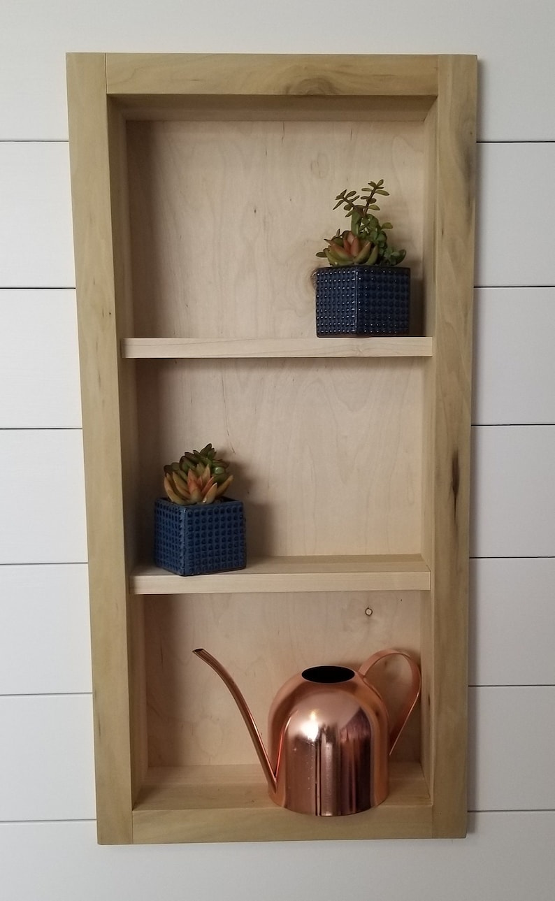 Pu&ograve; includere: Una mensola da parete in legno a tre livelli, con due piccoli vasi quadrati blu con succulente e un annaffiatoio in rame. La mensola &egrave; in legno chiaro ed &egrave; montata su una parete bianca.
