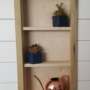 Pu&ograve; includere: Una mensola incassata in legno con tre ripiani. Due piccoli vasi quadrati blu navy con succulente sono sui due ripiani superiori. Un annaffiatoio in rame &egrave; sul ripiano inferiore.