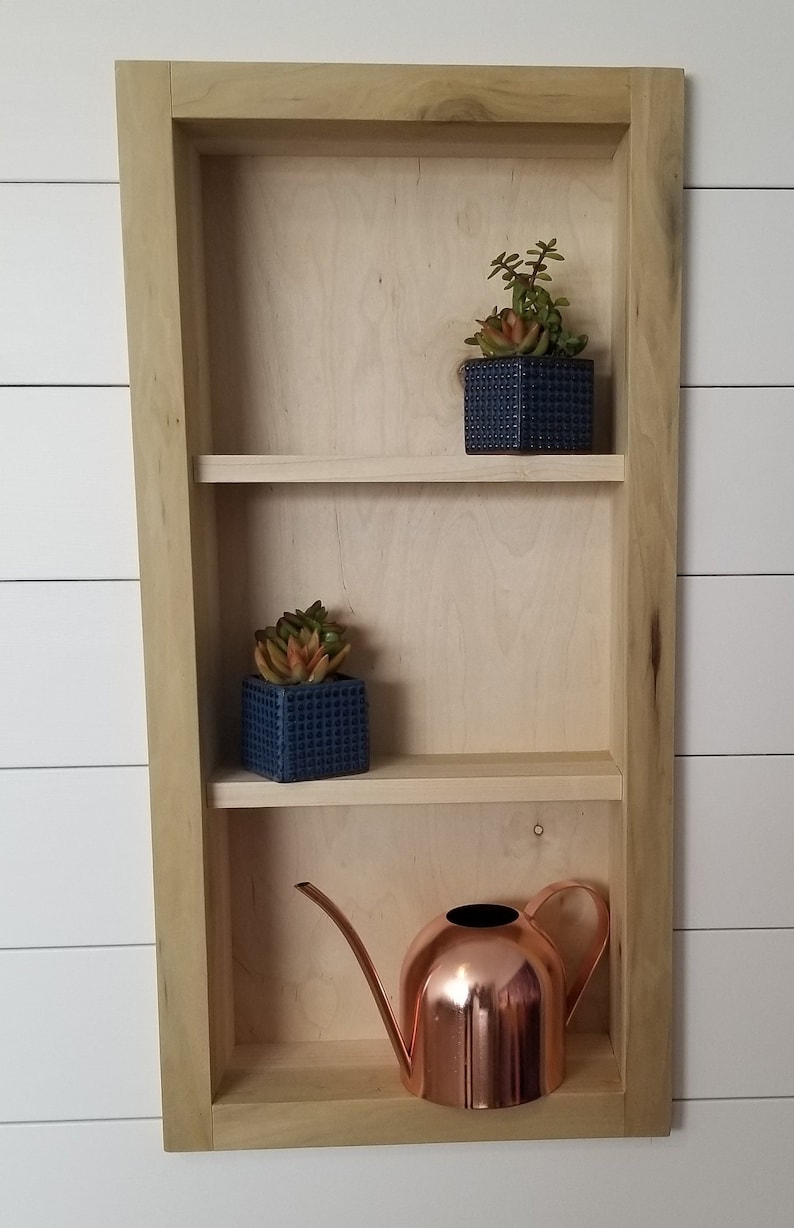 Pu&ograve; includere: Una mensola incassata in legno con tre ripiani. Due piccoli vasi quadrati blu navy con succulente sono sui due ripiani superiori. Un annaffiatoio in rame &egrave; sul ripiano inferiore.
