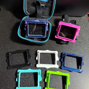 Puede incluir: Una colección de accesorios para ordenadores de buceo, que incluye un estuche de transporte azul y negro, dos ordenadores de buceo en fundas rosas y azules, y seis marcos protectores de colores en negro, blanco, azul, turquesa y verde. La palabra "WATER" está impresa en el estuche.