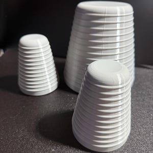 May include: Three white, ribbed, conical objects of varying sizes are arranged on a dark surface. The objects have a series of horizontal ridges, giving them a textured appearance. The largest object is approximately 5 inches tall.