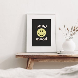 Może przedstawiać: Wydruk w ramie z czarnym tłem i białym tekstem, kt&oacute;ry brzmi "good mood" z ż&oacute;łtą buźką uśmiechniętą między słowami. Wydruk stoi na drewnianej p&oacute;łce z białym wazonen i suszonymi kwiatami.