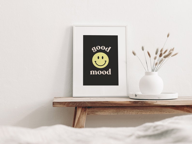 Może przedstawiać: Wydruk w ramie z czarnym tłem i białym tekstem, kt&oacute;ry brzmi "good mood" z ż&oacute;łtą buźką uśmiechniętą między słowami. Wydruk stoi na drewnianej p&oacute;łce z białym wazonen i suszonymi kwiatami.