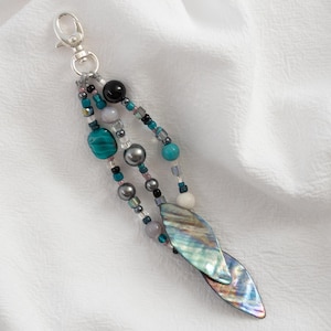 May include: A beaded keychain with a silver clasp. The keychain features strands of beads in teal, gray, black, and iridescent colors. Two iridescent shell-shaped charms dangle at the bottom, adding a touch of elegance to this accessory.