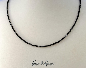 May include: A delicate necklace composed of small, faceted black beads strung together in a single strand. The necklace is displayed on a neutral-toned bust. Below the necklace, the text Hue & Haze is printed in a dark, cursive script font.