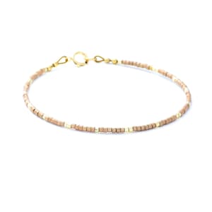 May include: A delicate bracelet featuring a repeating pattern of small, matte tan cylindrical beads interspersed with tiny, polished gold-tone metallic spacers. The piece is finished with a gold-tone metal spring ring clasp and jump ring.