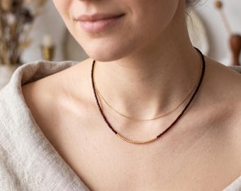 May include: A layered necklace set consisting of one thin gold chain and one beaded necklace featuring small dark red beads with a central section of tiny gold-toned beads. The necklaces are worn around the neck against a light beige fabric.