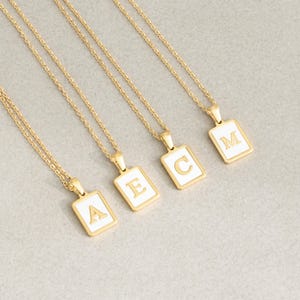 Puede incluir: Collares dorados con colgantes rectangulares con las letras A, E, C y M. Cada colgante tiene un fondo de esmalte blanco y una letra dorada. Los collares se muestran sobre una superficie gris claro.