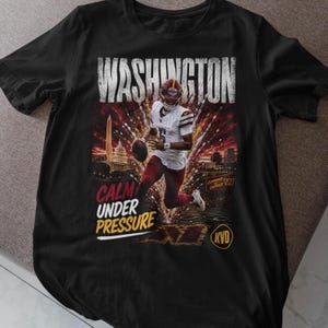 May include: A black short-sleeve t-shirt featuring a graphic of a football player in a white jersey and burgundy trousers. The text reads WASHINGTON, CALM UNDER PRESSURE, COMMAND DISTRICT, and KVD. The background shows a city skyline at sunset.