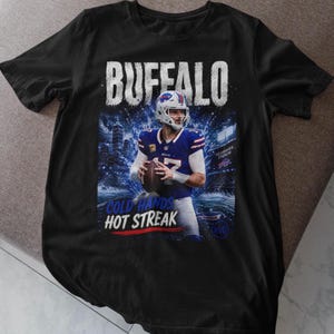 Puede incluir: Una camiseta negra con un estampado gráfico de un jugador de fútbol americano con uniforme azul y blanco sosteniendo un balón. El texto BUFFALO aparece en la parte superior, con COLD HANDS HOT STREAK impreso en blanco y azul bajo el jugador con un estadio.