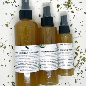 May include: Three amber glass bottles with spray tops, labelled "Rosemary-Moringa Hair Growth Tonic" and "Mary-Moringa Hair Growth Tonic". The bottles are on a white surface with green leaves scattered around them.