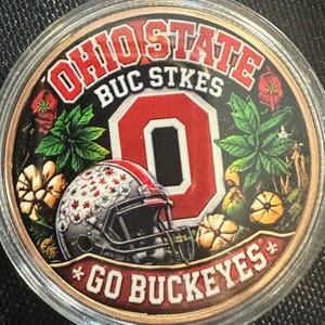 May include: A circular commemorative coin featuring the text OHIO STATE BUC STKES and GO BUCKEYES. The design includes a large red letter O, a football helmet with leaf patterns, and botanical illustrations of leaves and buckeye nuts.