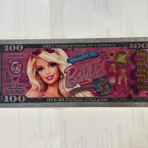 May include: A rectangular novelty item resembling a one hundred dollar bill with a pink background featuring a Barbie character illustration, the text Barbie, Chocolate Remember Me Game, and the number 100 in the corners with a metallic silver border.