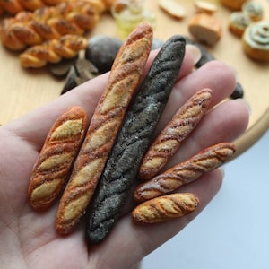 Puede incluir: Una colección de seis panes en miniatura hechos a mano, que incluyen una baguette oscura, una baguette blanca y cuatro panes más pequeños. Los panes están hechos de arcilla polimérica y están detallados de forma realista.