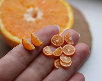 May include: A close-up of miniature orange slices and a large orange half. The tiny slices are held in a hand, showcasing the detail of the orange's texture and color. The background features a blurred wooden surface.