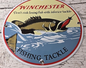 May include: Round metal sign with a red and white border featuring a fish jumping out of blue water. The sign reads "WINCHESTER FISHING TACKLE" in red and black text. The background is yellow.